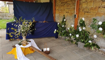 Im Vordergrund liegt ein Kreuz, es lehnt auf einem gelben Tuch, dahinter steht eine Blumenvase