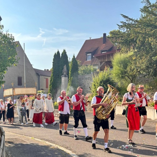 Blick auf die Dorfstraße. Man sieht die Musikkapelle beim Spielen und dahinter den Altardienst.